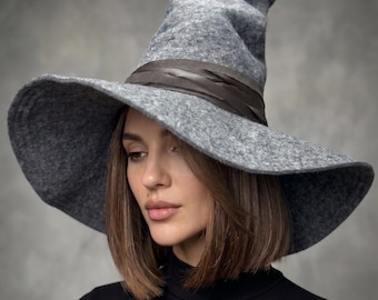 May include: A gray witch hat with a wide brim and a pointed crown. The hat is made of felt and has a dark brown ribbon around the base of the crown. The hat is worn by a person wearing a black turtleneck.