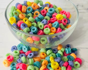 May include: A bowl filled with colorful cereal rings, with some spilling out onto a white surface. The cereal is a mix of green, yellow, orange, pink, and purple.