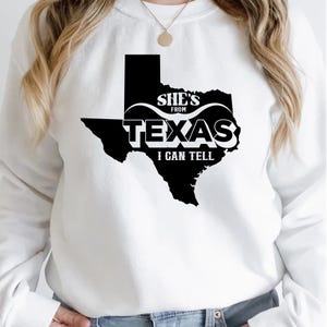 Studio style mockup of a woman seated indoors wearing a beige oversized sweatshirt printed with a bold black western design reading “She’s From Texas I Can Tell,” featuring a longhorn bull skull graphic integrated beneath the word Texas; model shown from shoulders to mid-thigh sitting on a bed with soft neutral background tones, light blue jeans visible, creating a cozy fall or winter fashion aesthetic;