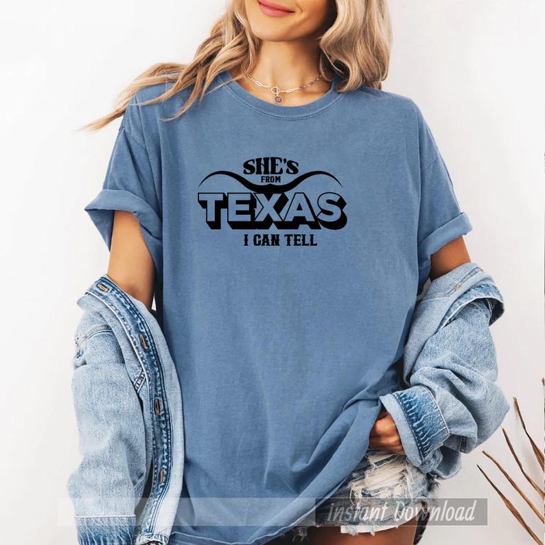 Studio style mockup of a woman seated indoors wearing a beige oversized sweatshirt printed with a bold black western design reading “She’s From Texas I Can Tell,” featuring a longhorn bull skull graphic integrated beneath the word Texas; model shown from shoulders to mid-thigh sitting on a bed with soft neutral background tones, light blue jeans visible, creating a cozy fall or winter fashion aesthetic;