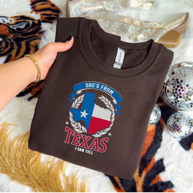 Studio style mockup of a woman seated indoors wearing a beige oversized sweatshirt printed with a bold black western design reading “She’s From Texas I Can Tell,” featuring a longhorn bull skull graphic integrated beneath the word Texas; model shown from shoulders to mid-thigh sitting on a bed with soft neutral background tones, light blue jeans visible, creating a cozy fall or winter fashion aesthetic;