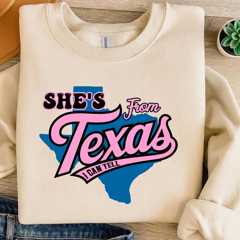 Studio style mockup of a woman seated indoors wearing a beige oversized sweatshirt printed with a bold black western design reading “She’s From Texas I Can Tell,” featuring a longhorn bull skull graphic integrated beneath the word Texas; model shown from shoulders to mid-thigh sitting on a bed with soft neutral background tones, light blue jeans visible, creating a cozy fall or winter fashion aesthetic;