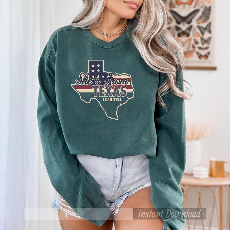Studio style mockup of a woman seated indoors wearing a beige oversized sweatshirt printed with a bold black western design reading “She’s From Texas I Can Tell,” featuring a longhorn bull skull graphic integrated beneath the word Texas; model shown from shoulders to mid-thigh sitting on a bed with soft neutral background tones, light blue jeans visible, creating a cozy fall or winter fashion aesthetic;