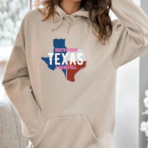 Studio style mockup of a woman seated indoors wearing a beige oversized sweatshirt printed with a bold black western design reading “She’s From Texas I Can Tell,” featuring a longhorn bull skull graphic integrated beneath the word Texas; model shown from shoulders to mid-thigh sitting on a bed with soft neutral background tones, light blue jeans visible, creating a cozy fall or winter fashion aesthetic;
