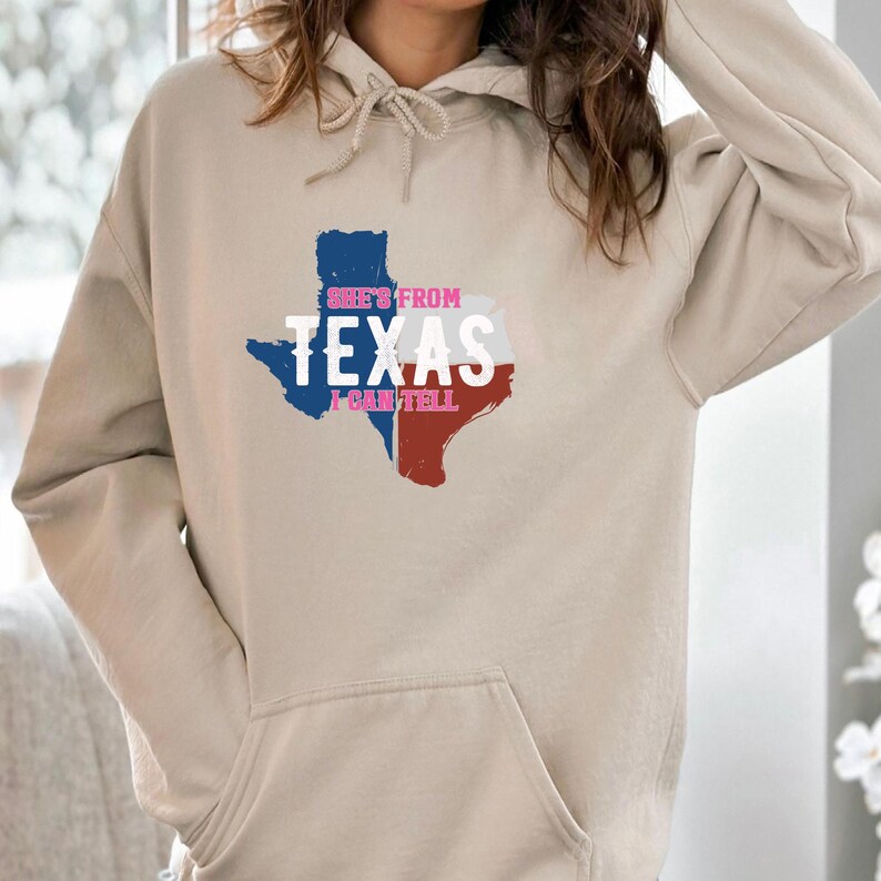 Studio style mockup of a woman seated indoors wearing a beige oversized sweatshirt printed with a bold black western design reading “She’s From Texas I Can Tell,” featuring a longhorn bull skull graphic integrated beneath the word Texas; model shown from shoulders to mid-thigh sitting on a bed with soft neutral background tones, light blue jeans visible, creating a cozy fall or winter fashion aesthetic;
