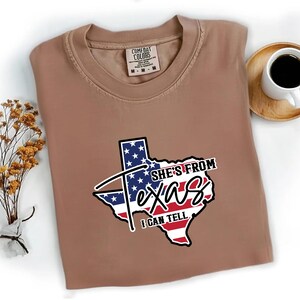 Studio style mockup of a woman seated indoors wearing a beige oversized sweatshirt printed with a bold black western design reading “She’s From Texas I Can Tell,” featuring a longhorn bull skull graphic integrated beneath the word Texas; model shown from shoulders to mid-thigh sitting on a bed with soft neutral background tones, light blue jeans visible, creating a cozy fall or winter fashion aesthetic;