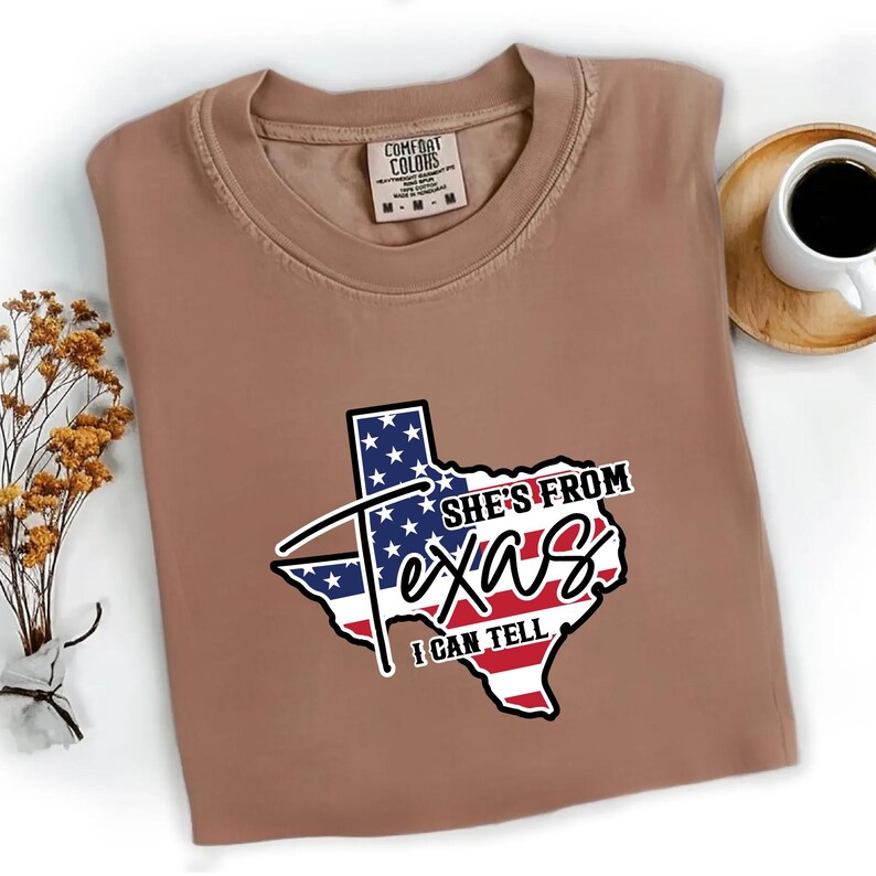 Studio style mockup of a woman seated indoors wearing a beige oversized sweatshirt printed with a bold black western design reading “She’s From Texas I Can Tell,” featuring a longhorn bull skull graphic integrated beneath the word Texas; model shown from shoulders to mid-thigh sitting on a bed with soft neutral background tones, light blue jeans visible, creating a cozy fall or winter fashion aesthetic;