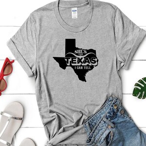 以下が含まれることがあります： 「She's from Texas I can tell」の黒い文字とテキサスの形がプリントされたグレーのTシャツ。Tシャツは折りたたまれており、赤いサングラスとシルバーのサンダルが見えます。Tシャツは綿製です。