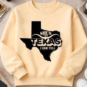 Studio style mockup of a woman seated indoors wearing a beige oversized sweatshirt printed with a bold black western design reading “She’s From Texas I Can Tell,” featuring a longhorn bull skull graphic integrated beneath the word Texas; model shown from shoulders to mid-thigh sitting on a bed with soft neutral background tones, light blue jeans visible, creating a cozy fall or winter fashion aesthetic;