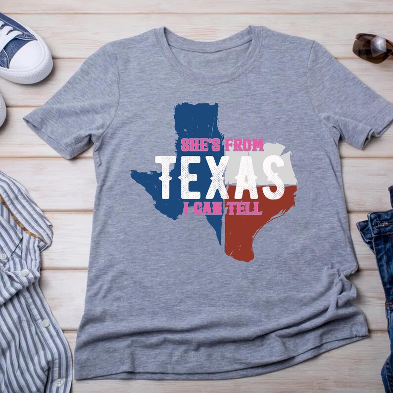 Studio style mockup of a woman seated indoors wearing a beige oversized sweatshirt printed with a bold black western design reading “She’s From Texas I Can Tell,” featuring a longhorn bull skull graphic integrated beneath the word Texas; model shown from shoulders to mid-thigh sitting on a bed with soft neutral background tones, light blue jeans visible, creating a cozy fall or winter fashion aesthetic;