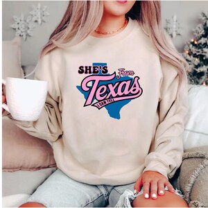 Studio style mockup of a woman seated indoors wearing a beige oversized sweatshirt printed with a bold black western design reading “She’s From Texas I Can Tell,” featuring a longhorn bull skull graphic integrated beneath the word Texas; model shown from shoulders to mid-thigh sitting on a bed with soft neutral background tones, light blue jeans visible, creating a cozy fall or winter fashion aesthetic;