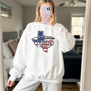 Lifestyle mockup of a young woman wearing a light gray knotted t-shirt featuring a Texas state outline graphic filled with an American flag design and bold script text reading “She’s From Texas I Can Tell,” printed in black, red, white, and blue on the center chest; model shown from shoulders to waist with soft natural makeup and brown hair, standing near a wooden bench with green houseplants in the
