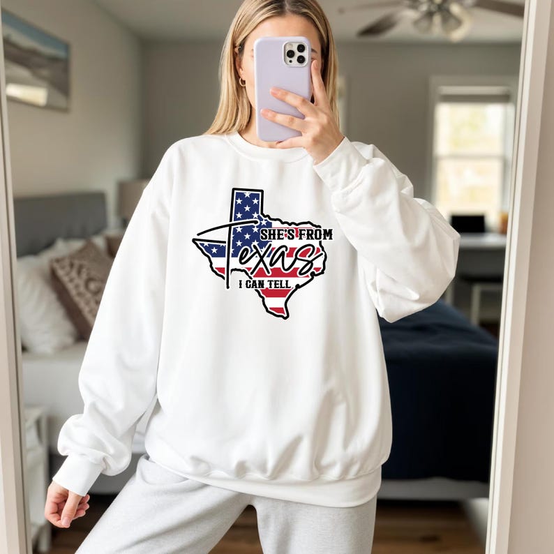 Lifestyle mockup of a young woman wearing a light gray knotted t-shirt featuring a Texas state outline graphic filled with an American flag design and bold script text reading “She’s From Texas I Can Tell,” printed in black, red, white, and blue on the center chest; model shown from shoulders to waist with soft natural makeup and brown hair, standing near a wooden bench with green houseplants in the
