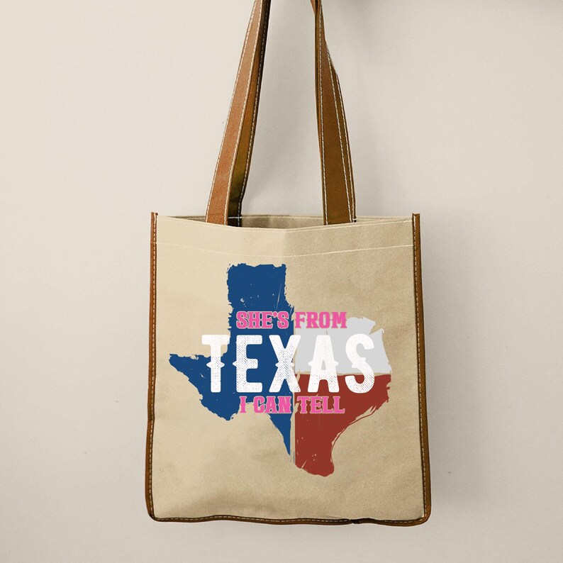 Flat lay mockup of a TOTE BAG displayed on a WHITE surface with minimal decor accents, featuring a bold black Texas state silhouette graphic with layered western typography reading “She’s From Texas I Can Tell” across the center; design includes curved accent lines and strong block lettering optimized for vinyl cutting, heat transfer, or screen printing; shirt styled with a delicate gold necklace placed above the collar and subtle dried botanical elements at the side