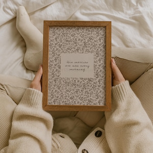 May include: A rectangular wooden frame containing a print with a small-scale gray floral pattern on a white background. Centered on the print is a white rectangular label featuring the black cursive text, His mercies are new every morning.