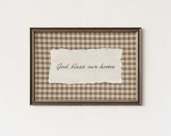 May include: A rectangular wall art piece in a dark frame featuring a beige and brown gingham pattern background. Centered is a piece of off-white paper with torn edges displaying the dark brown printed text, God bless our home.