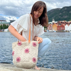 Op de afbeelding: Een vrouw houdt een gehaakte tote bag vast met roze bloemen. De tas heeft bruine leren hengsels. De vrouw draagt een wit shirt en een blauwe jeans. Ze staat voor een waterpartij met kleurrijke gebouwen op de achtergrond.