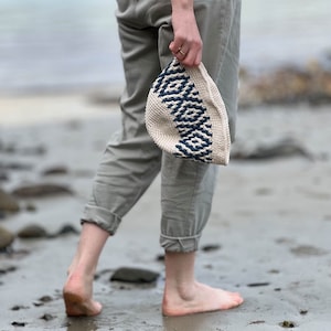 Può includere: Una persona che cammina su una spiaggia tenendo un cappello all'uncinetto bianco e blu.