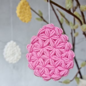 Könnte beinhalten: Ein rosa gehäkeltes Osterei-Anhänger, der an einem Zweig hängt. Das Ei ist aus Garn gefertigt und hat ein blumenartiges Muster.