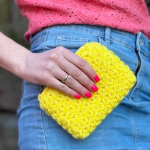 May include: A bright yellow crocheted coin purse with a flower pattern. The purse is held in a hand with pink nail polish.