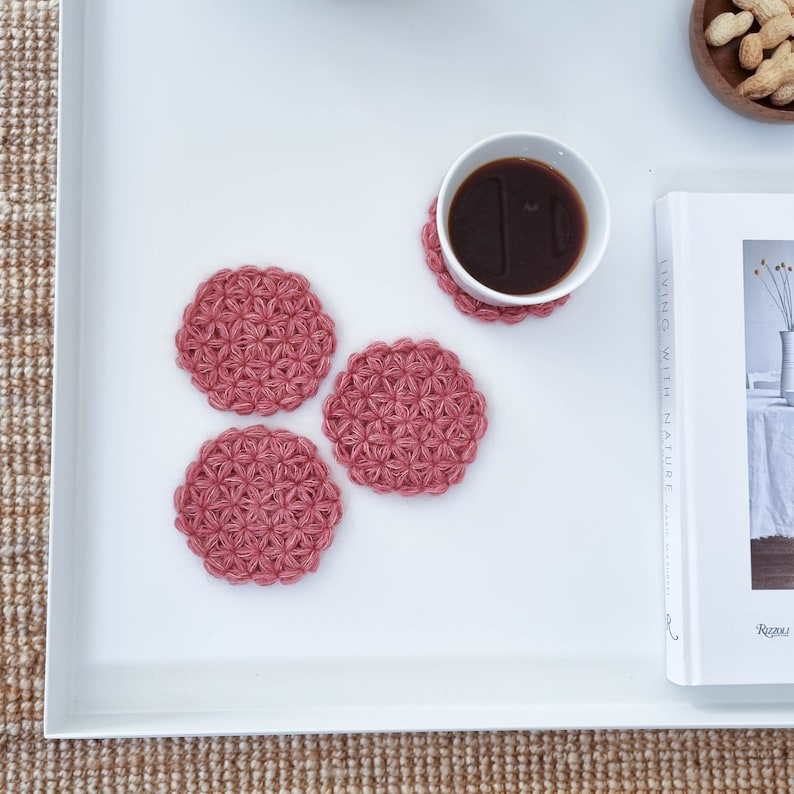 Puede incluir: Cuatro posavasos de crochet rosa con un patr&oacute;n de flores, sobre una superficie blanca con una taza de caf&eacute; y un libro titulado "Vivir con la naturaleza" a un lado.