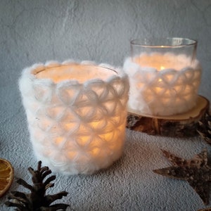 May include: Two white crocheted candle holders with a glass votive inside each. The candle holders are lit and casting a warm glow. The candle holders are sitting on a wooden surface.
