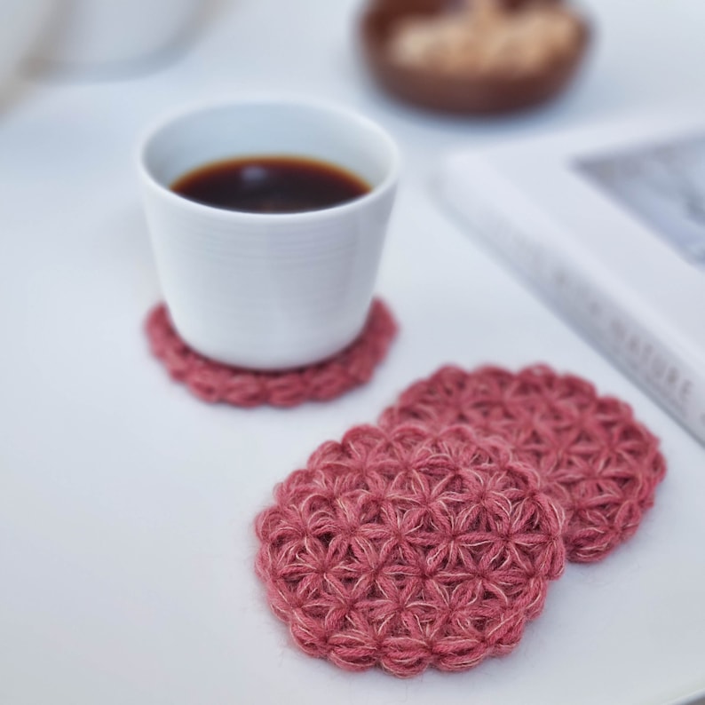 Puede incluir: Dos posavasos de crochet rosa con un patr&oacute;n de flores. Una taza de cer&aacute;mica blanca con caf&eacute; se encuentra sobre uno de los posavasos.