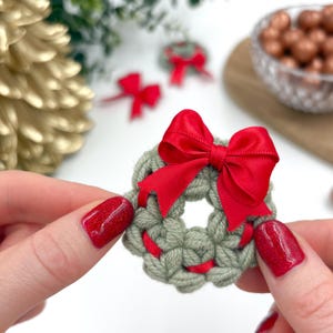 Könnte beinhalten: Ein kleiner, handgefertigter Häkelkranz mit einer roten Satinschleife. Der Kranz hat eine gedämpfte grüne Farbe mit roten Akzenten. Das Bild zeigt den Kranz zwischen zwei Händen, mit anderen Kränzen und dekorativen Elementen im Hintergrund.