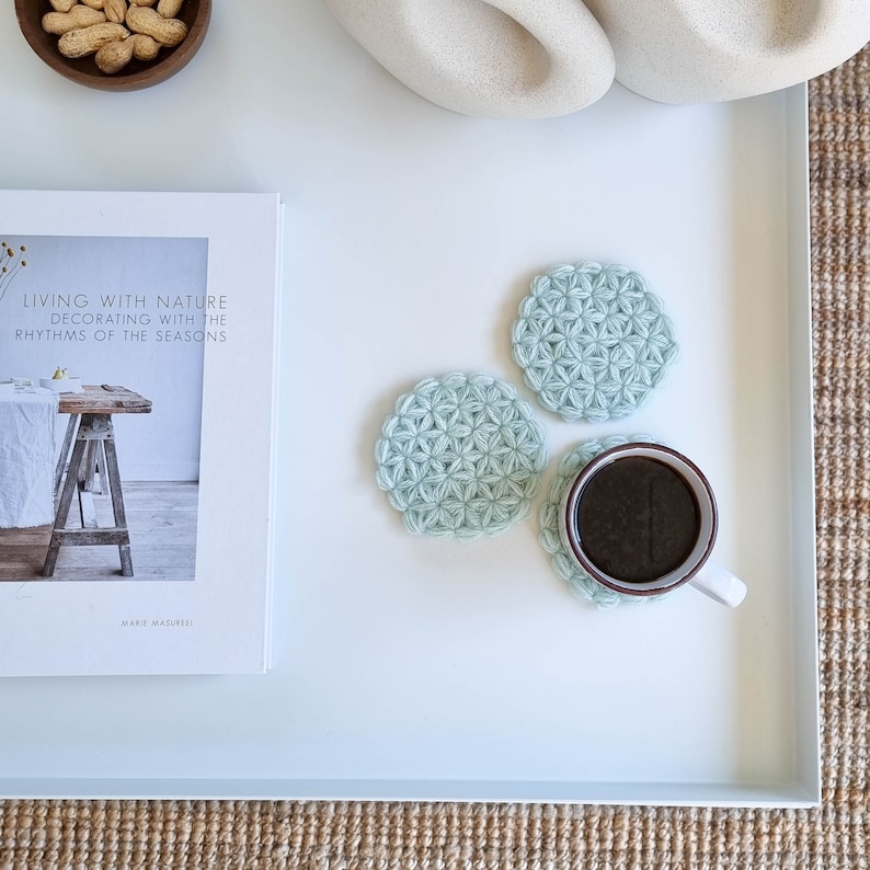 Puede incluir: Tres posavasos de crochet azul claro con un patr&oacute;n de estrella, uno de los cuales sostiene una taza de caf&eacute;. Los posavasos est&aacute;n sobre una superficie blanca.