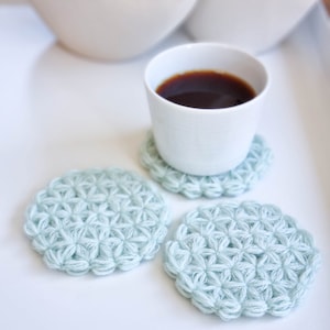 Puede incluir: Tres posavasos de ganchillo de color azul claro con un patr&oacute;n de estrella. Una taza blanca con caf&eacute; se encuentra sobre uno de los posavasos.