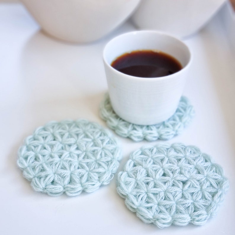 Puede incluir: Tres posavasos de ganchillo de color azul claro con un patr&oacute;n de estrella. Una taza blanca con caf&eacute; se encuentra sobre uno de los posavasos.