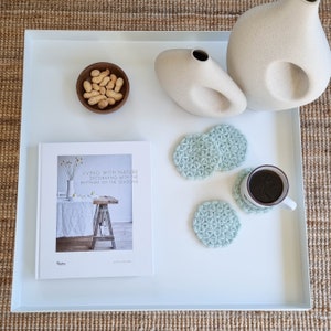 Puede incluir: Una bandeja blanca con un libro titulado "Vivir con la naturaleza Decorar con los ritmos de las estaciones" de Marie Olsson Nylander, un cuenco de cacahuetes, dos jarrones de cer&aacute;mica blanca y cuatro posavasos de crochet azul claro. Un posavasos tiene una taza de caf&eacute; encima.