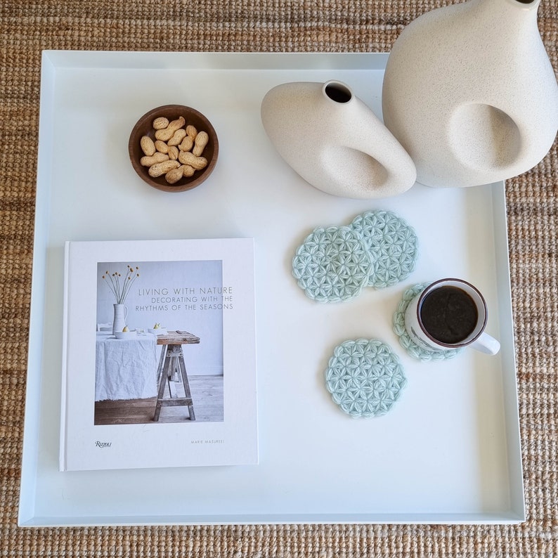 Puede incluir: Una bandeja blanca con un libro titulado "Vivir con la naturaleza Decorar con los ritmos de las estaciones" de Marie Olsson Nylander, un cuenco de cacahuetes, dos jarrones de cer&aacute;mica blanca y cuatro posavasos de crochet azul claro. Un posavasos tiene una taza de caf&eacute; encima.