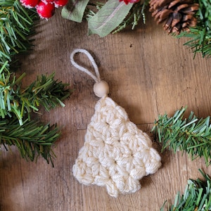 Puede incluir: Un adorno de &aacute;rbol de Navidad de crochet blanco con detalles de hilo dorado y una cuenta de madera en la parte superior.