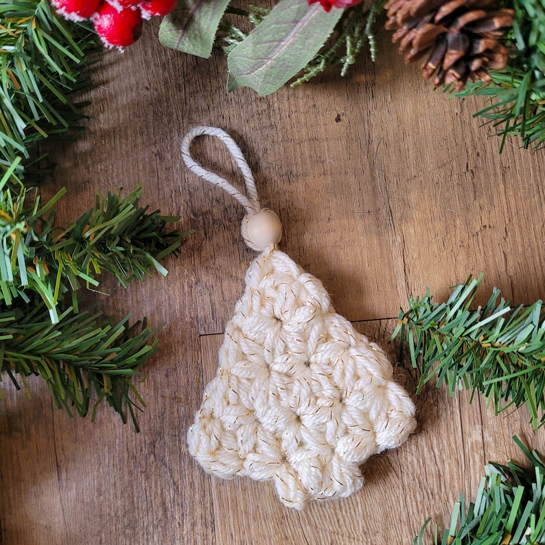 Puede incluir: Un adorno de &aacute;rbol de Navidad de crochet blanco con detalles de hilo dorado y una cuenta de madera en la parte superior.