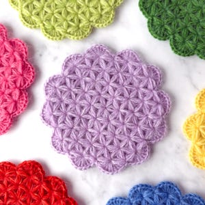 May include: A collection of colorful, handmade crochet coasters in various shades, including purple, green, pink, red, yellow, and blue. Each coaster features a flower-like shape with a detailed, textured stitch pattern. The coasters are arranged on a white surface.