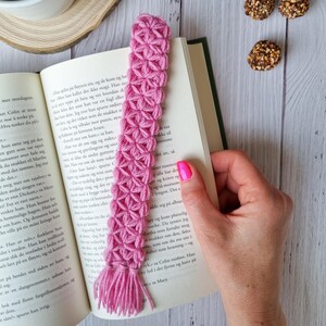 Può includere: Un segnalibro rosa all'uncinetto con un fiocco. Il segnalibro è realizzato con un motivo floreale ripetitivo.