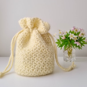 May include: A crocheted drawstring bag made of cream-coloured yarn with a flower pattern. The bag is sitting on a white surface next to a vase of white and pink flowers.