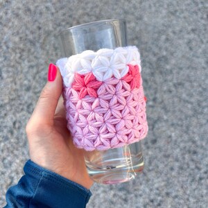 Op de afbeelding: Een helder glas gevuld met vloeistof is omhuld in een handgemaakte, gehaakte hoes. De hoes heeft een bloemenpatroon in een verloop van roze en witte tinten. De hoes wordt vastgehouden door een hand met felroze nagellak.