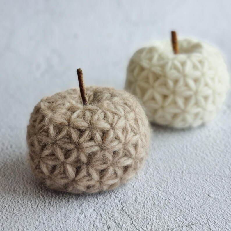May include: Two crocheted apples, one brown and one white, with a textured, flower-like pattern. The apples are sitting on a light gray surface.