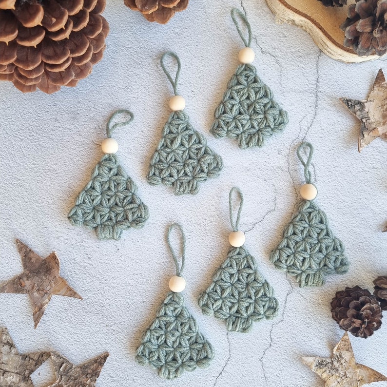 May include: Six crocheted green Christmas tree ornaments with wooden beads. The ornaments are hanging from white string on a light gray background.