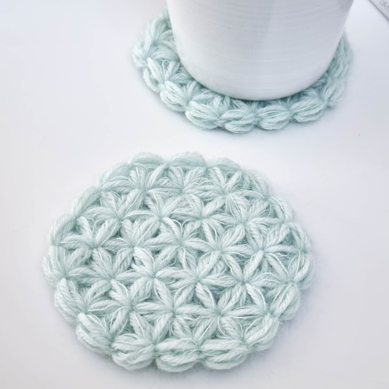 Puede incluir: Dos posavasos de ganchillo de color azul claro con un patr&oacute;n de flores. Los posavasos son redondos y tienen una textura en relieve.