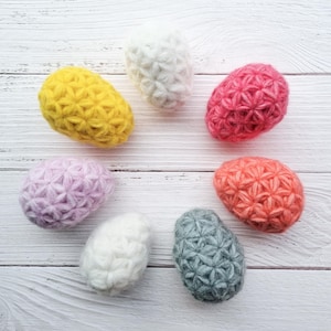 Pode incluir: Uma coleção de sete objetos decorativos em forma de ovo coloridos. Os ovos são em tons de amarelo, branco, rosa, roxo, laranja e cinza. Cada ovo tem um padrão texturizado em forma de estrela em sua superfície.