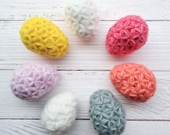 May include: Seven decorative, egg-shaped objects in various colours. The eggs are yellow, white, pink, purple, orange, and grey. Each egg has a textured, star-like pattern across its surface.