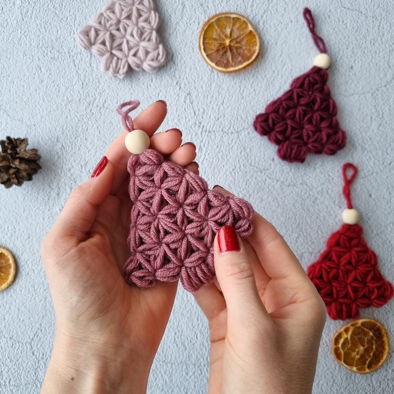 Puede incluir: Primer plano de las manos de una persona sosteniendo un adorno de &aacute;rbol de Navidad de crochet rosa. El adorno est&aacute; hecho de hilo y tiene una cuenta de madera en la parte superior.