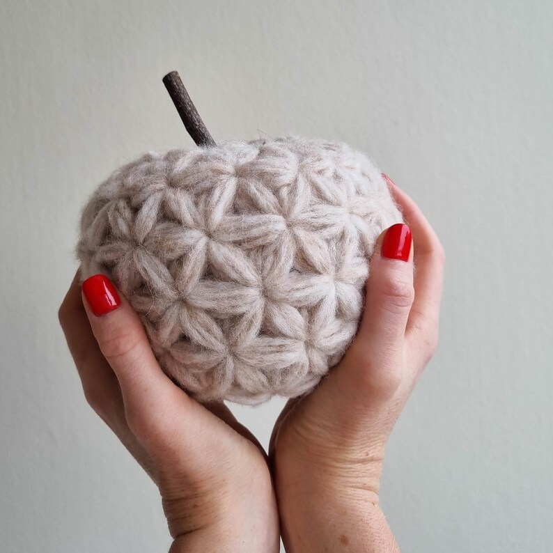 May include: A beige crocheted apple with a starburst pattern, held in hands with red nail polish.