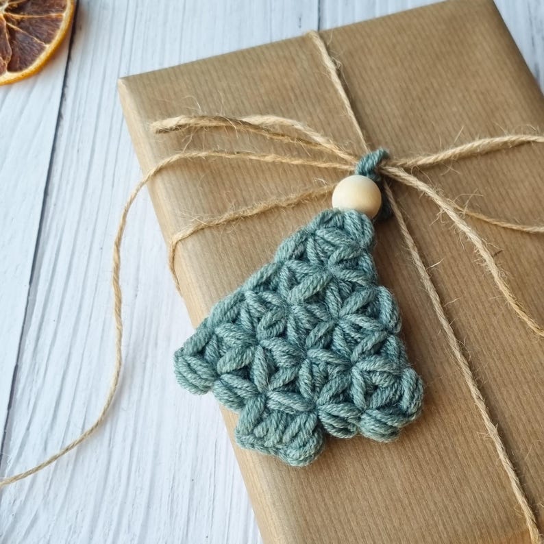 Puede incluir: Un adorno de &aacute;rbol de Navidad de crochet de color verde azulado con una cuenta de madera en la parte superior, atado con una cuerda a una caja de regalo marr&oacute;n.