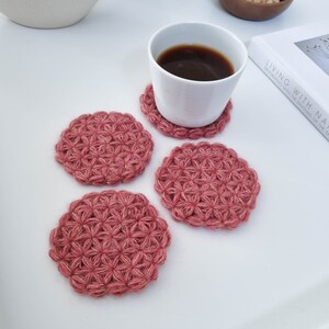 Puede incluir: Cuatro posavasos de ganchillo en color rosa polvoriento con un patr&oacute;n de flores. Los posavasos est&aacute;n sobre una superficie blanca con una taza de caf&eacute; blanca y un libro.