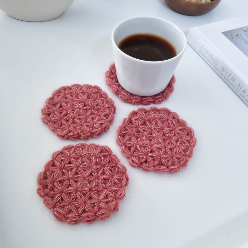 Puede incluir: Cuatro posavasos de ganchillo en color rosa polvoriento con un patr&oacute;n de flores. Los posavasos est&aacute;n sobre una superficie blanca con una taza de caf&eacute; blanca y un libro.