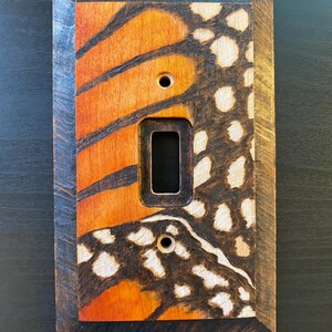 May include: Wooden light switch cover featuring a monarch butterfly wing design. The cover has a rectangular opening for the switch and two holes for screws. The butterfly wing design is orange, black, and white, with a dark brown wooden frame.
